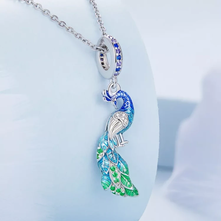 Wiszący charms Paw z zielono-niebieskimi zdobieniami i połyskującymi cyrkoniami, pokazany w eleganckiej aranżacji produktowej. Oryginalna zawieszka Amélioré z motywem pawia, kompatybilna z bransoletkami modułowymi Pandora i Apart.