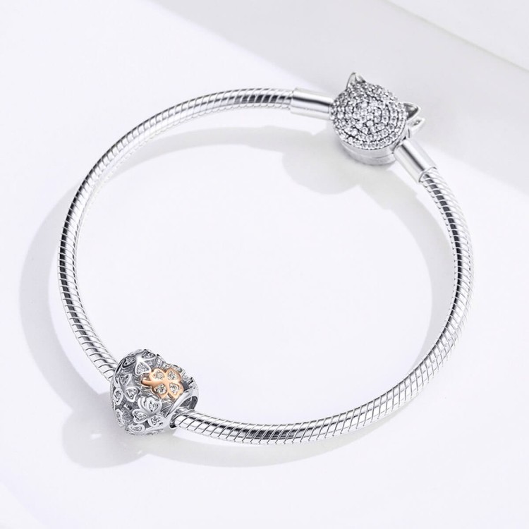 Ażurowy charms Serce Koniczynki prezentowany na bransoletce modułowej. Zawieszka Amélioré ozdobiona białymi cyrkoniami oraz czterolistną koniczynką w kolorze rose gold. Symboliczny charms łączący motyw miłości i szczęścia.