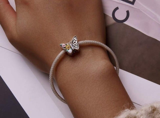 Charms Kwiecisty Motyl z ażurowym srebrzystym skrzydłem, kolorowymi kwiatami oraz błyszczącymi białymi cyrkoniami. Ozdobna zawieszka Amélioré wyeksponowana na bransoletce.